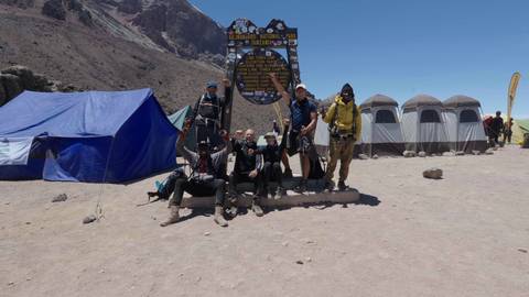 Trekking team poses triumphantly beneath the Lava Tower Camp sign on dusty slopes of Kilimanjaro with tents behind.