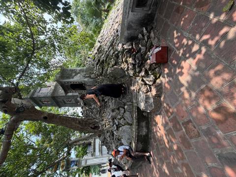 Visitor climbs rocky mound toward a small stone shrine shaded by large trees.