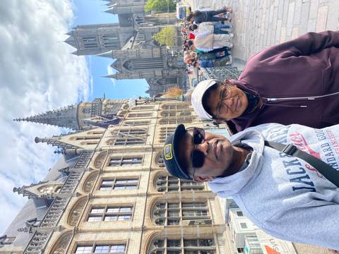 An older couple poses on a stone bridge in a historic European city, with ornate gothic buildings and a tall church spire behind them under a partly cloudy sky.