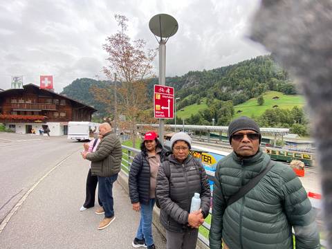 Five travellers in warm jackets stand beside an alpine road with Swiss directional signs, rustic chalets and green mountains in the background.
