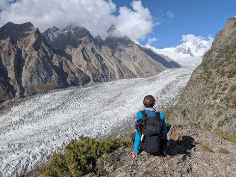 A hiker with a backpack looks over a vast white glacier snaking through dramatic jagged mountains under a bright blue sky.