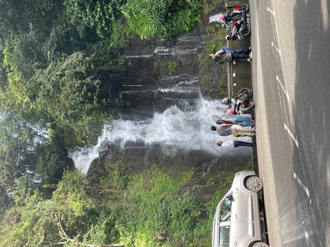 Travelers stop beside parked motorcycles to admire a roadside waterfall tumbling down a mossy cliff.