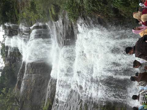 Crowds gather at the base of a tall, multi-drop waterfall plunging down a rocky cliff.