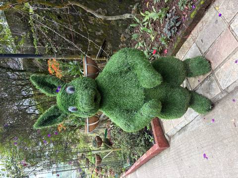 Whimsical rabbit-shaped topiary with googly eyes stands on a garden path surrounded by flowers.