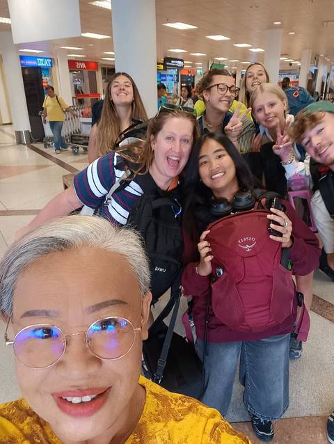 Group selfie of excited travelers wearing backpacks inside an airport terminal.