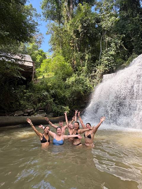 Laughing travelers stand waist-deep in a jungle waterfall pool with arms raised.