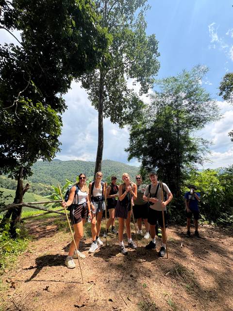 Hiking group pauses on a lush jungle trail with mountain scenery behind.