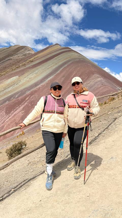 Two female hikers in matching fleece jackets smile for the camera on the colorful striped slope of Peru’s Rainbow Mountain trail.