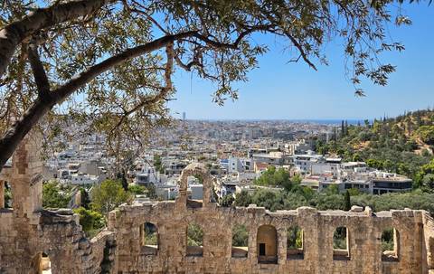 Framed by olive branches, Athens’ sprawling urban landscape meets the blue horizon beyond ancient stone ruins.