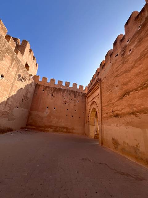 An open courtyard inside high crenellated walls, showing textured adobe surfaces lit by strong sunlight.