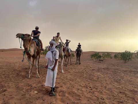 Tourists ride camels led by a Tuareg guide across reddish Sahara dunes at dusk.