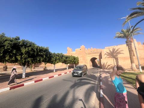 Street scene outside adobe walls with palms, a car, and casual pedestrians under bright sun.