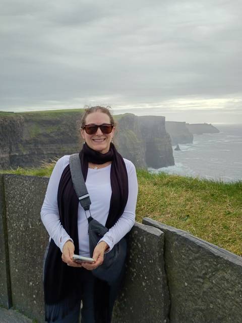 Smiling woman in sunglasses poses by a windswept grassy edge overlooking misty sea cliffs.