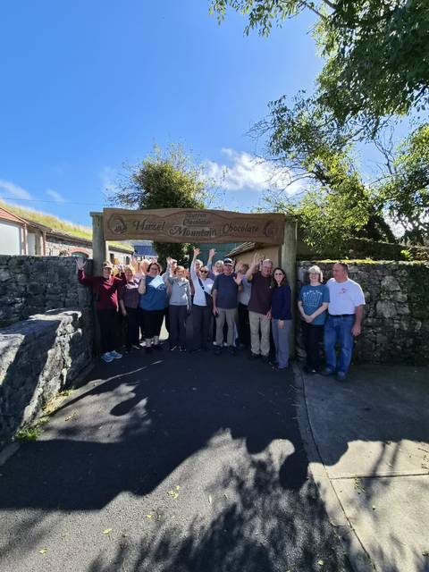 Tour group cheers under a wooden archway for Hazel Mountain Chocolate with stone walls and blue sky.