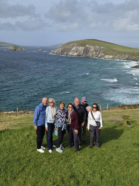 Small group of travellers stand on a grassy cliffside above a choppy blue sea and rugged coastline.