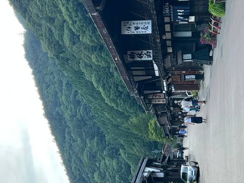 Quiet rural Japanese street lined with dark-wood Edo buildings and forested hills rising behind.