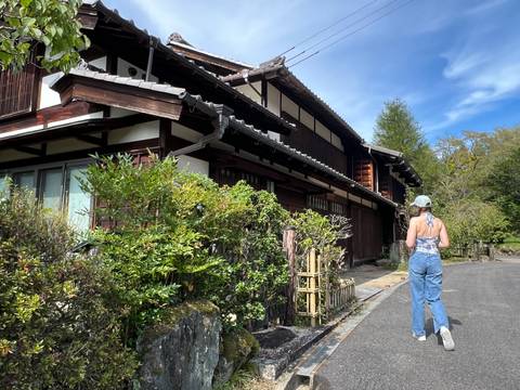Young traveller strolls past a traditional timber house framed by lush garden plants under blue sky.