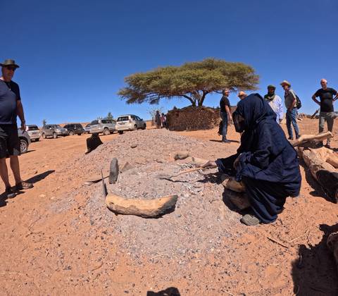 Berber guide in blue robe tending a traditional desert earth oven while travellers watch nearby vehicles