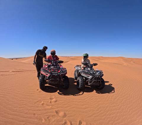 Two quad bikes with riders and a guide stopped on rolling orange sand dunes under a cloudless sky.
