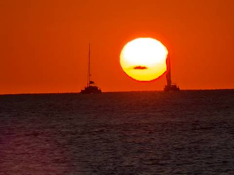 Vivid orange sunset over the sea with two sailboats silhouetted against a giant setting sun.