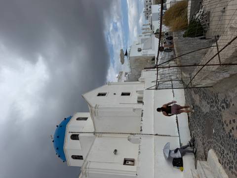 Whitewashed church with blue dome beneath dramatic grey clouds, tourist walking along a cobbled path.