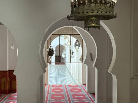 Elegant white arches frame a mosaic-tiled corridor and ornate hanging lanterns inside a Moroccan mosque or madrasa.