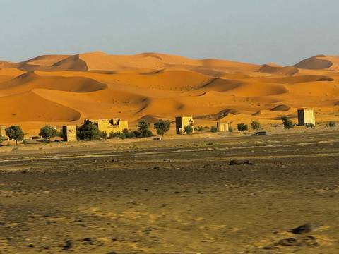 Small desert settlement framed by sweeping orange sand dunes under a hazy sky.