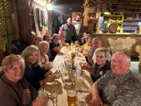 Happy tour group sharing drinks and dinner around a long rustic wooden table.