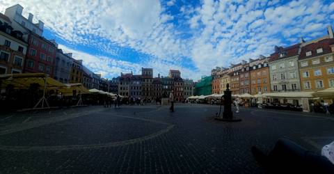 Wide cobblestone square in Warsaw Old Town surrounded by colorful historic facades under a patterned cloud sky.