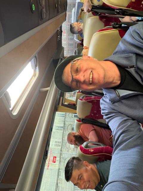 Smiling traveler takes a selfie inside a tour bus with maroon seats and city buildings outside the window.