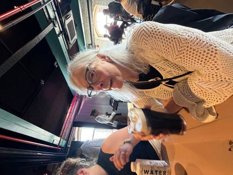 An older woman wearing glasses and a knitted cardigan enjoys a dark beer inside a lively pub with modern décor.
