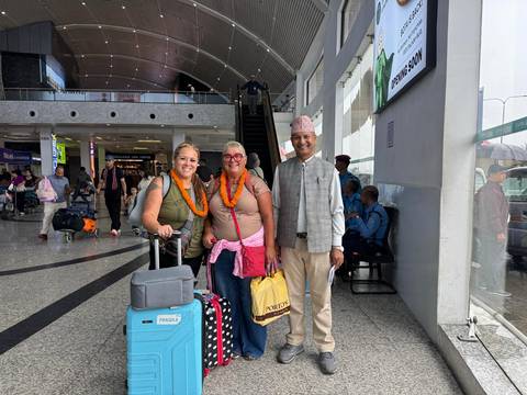 Two female travellers with luggage and marigold garlands stand with a smiling local guide inside an airport arrivals hall.