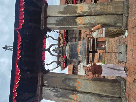 Two women pose on either side of a massive hanging bell in a historic courtyard temple with stone pillars and red trim.