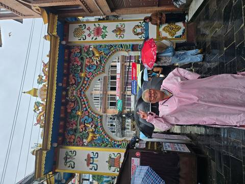 Woman in a pink rain poncho flashes a peace sign beneath an ornate colorful arch while others shelter under umbrellas.