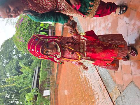 Young child dressed in elaborate red traditional costume with jewellery and makeup holds an adult's hand.