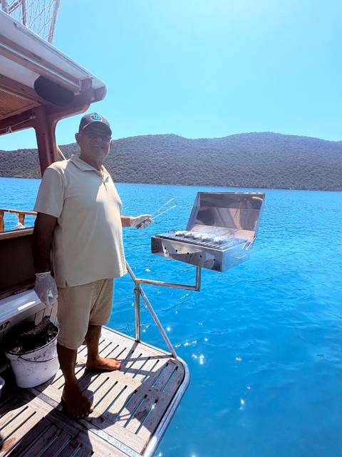 man grilling fish on stainless barbecue at back of boat with bright blue water
