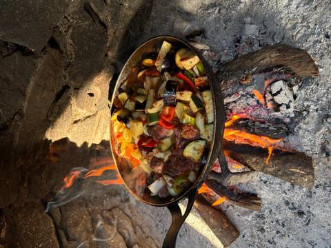 A cast-iron pan full of mixed vegetables and meat simmering over a wood fire beside a stone wall.