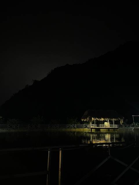 A dark night scene showing the silhouette of a forested mountain and a small thatched shelter lit below.