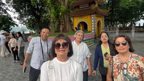 Selfie of travelers in front of a yellow pagoda-style shrine inside a leafy courtyard.