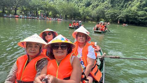 Visitors wearing life jackets and conical hats ride in a wooden rowboat on a green river.