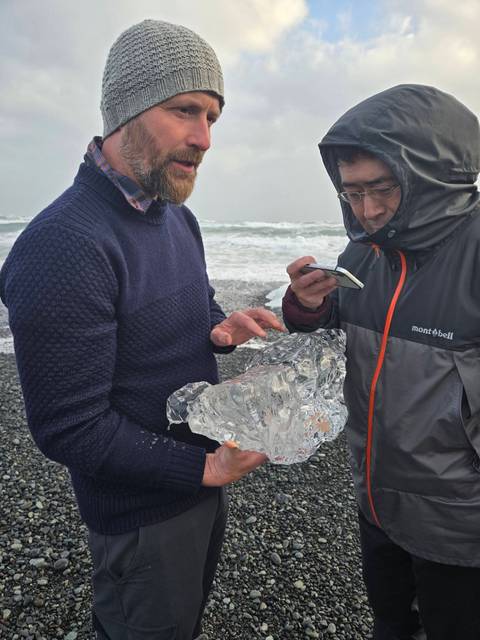 Travellers examine a clear ice chunk on a windswept volcanic beach near crashing surf.