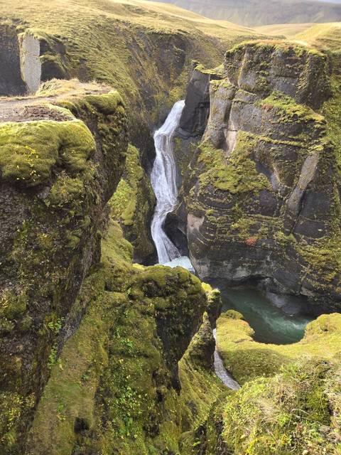 Narrow waterfall cascades through a moss-covered gorge into a turquoise pool deep within volcanic cliffs.
