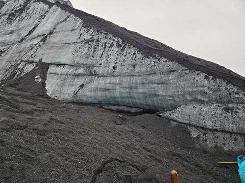 Stark face of a grey glacier with striations and dark volcanic ash layers under an overcast sky.