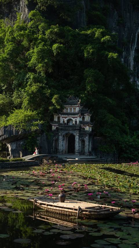 Ancient Vietnamese temple gate nestled in dense jungle cliffs with lone visitor in bright dress.