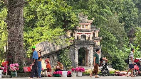 Local vendors sell pink lotus flowers near ornate jungle temple entrance in Vietnam.