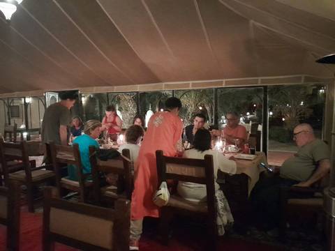 A large group of travellers sit around a long wooden table inside a canvas-roofed desert camp restaurant, dining by candlelight.
