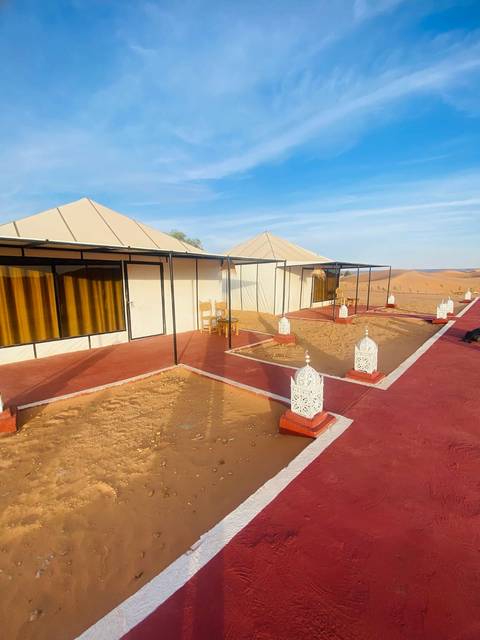 Rows of cream-coloured canvas tents with red walkways and decorative white lanterns stand in sandy dunes under a blue sky.