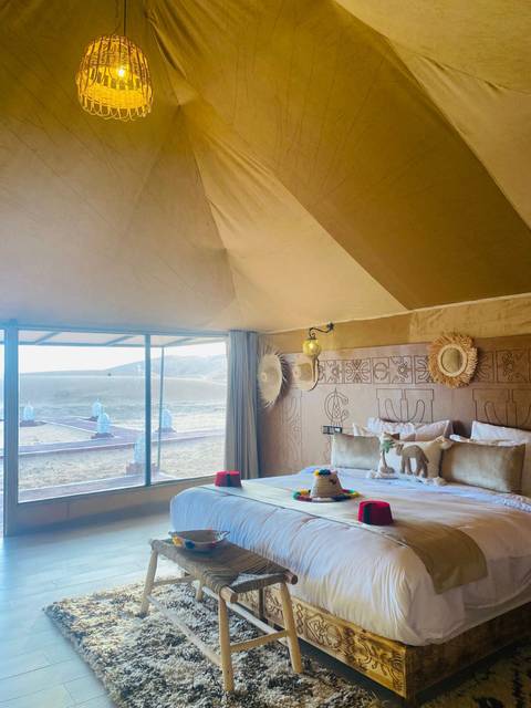 Stylish desert-camp bedroom with patterned pillows, woven decor and a large window looking onto rolling dunes.