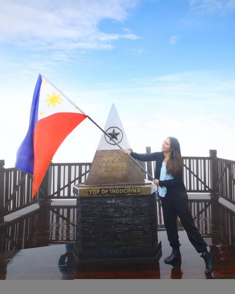 A smiling woman waves a national flag beside the Fansipan summit marker above the clouds.