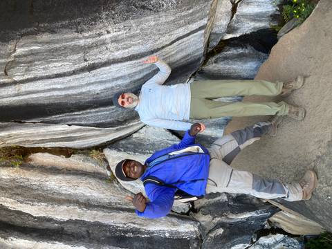 Two hikers posing thumbs-up beside dramatic striped rock face on mountain trail.